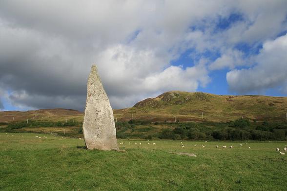 A good dramatic sky - blue showing through the clouds, a hill in sunshine, and in the foreground a very tall pointy standing stone also in sunshine. green grass, distant sheep.