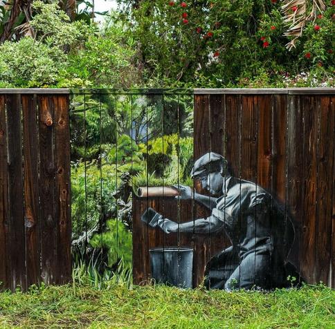 Streetart. On a dark brown wooden garden fence is painted mural of a man, who is sticking the fence. The man is painted in green-gray, he wears work pants, short shirt and flat cap. He is kneeling on the ground and using a brush to spread glue for a wallpaper on the fence. Two pieces of wallpaper can already be seen in the middle of the fence. They show exactly the background of the real fence. A wallpaper with the trees and bushes of the real garden. A great effect, because the fence picture was worked very realistically. Due to the fact that the figure is drawn in gray, the colorful drawing looks even more real and confuses the viewer.
The fence painting was painted during the Covid period in the artist's garden.