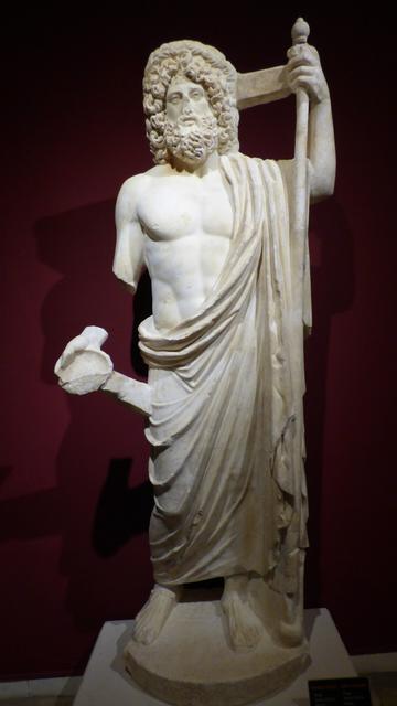 a roman statue of the God of Healing, Asclepius. He stands bare-chested, In his right hand he holds a bowl, his left arm is upraised holding his staff.
