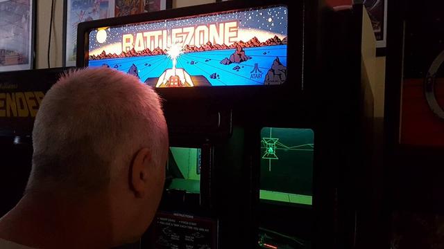 A person looks through the viewfinder of an original Atari BattleZone cabinet. On the screen is a tank in the player's sights. The marquee display is a brightly coloured space scene - a firing tank crosses a planet with the "BATTLEZONE" logo across the top.