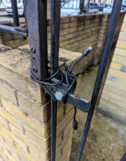 Close up of the latch of a closed gate and a brick wall. The gate is held shut by a mini USB to USB A wire wrapped repeatedly around both sides of gate and fence.