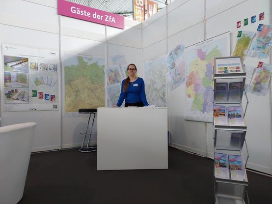 Weißer Messestand mit farbigen Landkarten an den Wänden. In der Mitte ein weißer Tresen. Dahinter steht eine Frau mit blauem Oberteil. Obe drüber der Text: Gäste der ZfA