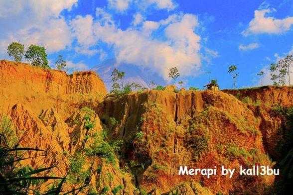 Vulcan Merapi behind yellow brown steep hill and against cloud blue sky.