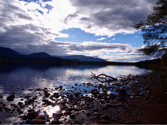 Photo towards the end of the day, muted shades of blue, grey, white with silhouetted hills and trees. Grey white cloud gathers in the left of the shot with bright patches of white and blue where the sun is covered. Hills mid section and Loch Morlich below, reflecting the clouds. A rocky foreshore, a tree branch silhouetted in the water on the shoreline, a tree in leaf