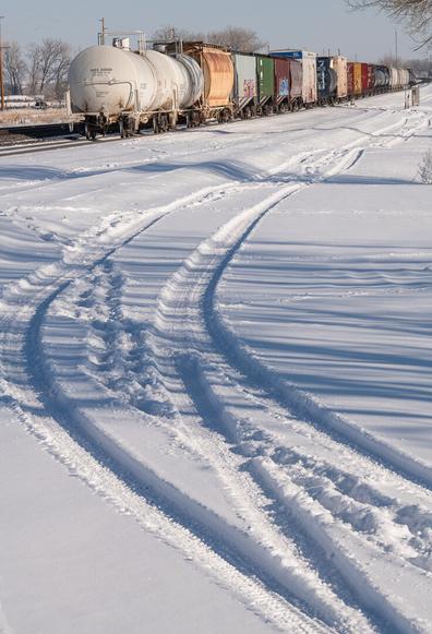 A line of colourful and rather rusty railway trucks stand idle on a very minor rail track. In the foreground there is deep snow with vehicle tracks in it visible