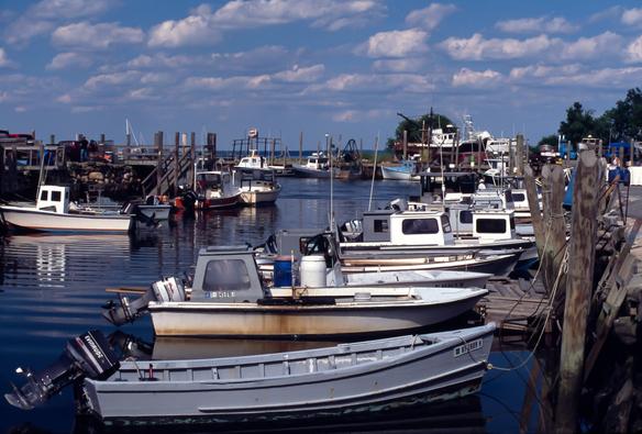 A color photograph on slide film. It shows small boats, mostly with outboards, tied up along two docks, back to back with a channel of very blue water between them. A bright summer day, there are puffy white clouds in the blue sky. In the background, sitting on green grass on the opposite shore, a couple of boats have been hauled out at a local shipyard. A Texaco fuel sign is visible nearby.