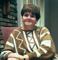 Photo of Invisible Disability Rights Hero, Elaine Wilson wearing a light brown and white sweater with straight and angled decorative lines.  Earings hang just below her short dark hair as she looks confident and content with hands resting on one arm of the chair.