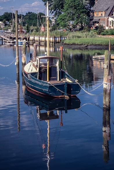 A photograph taken on a sunny summer day of a small wooden boat (inboard, visible rudder), with a dark blue painted hull, a stained wood cabin deck, and a white painted foredeck. There’s a small cabin area forward, and  a mast with no boom above the small pilot house and wheel in the center. 

The boat is moored in calm blue water, and a reflection of the boat is fully visible.  Wooden pilings surround it, and in the background is a house on land covered in bright green grass.
