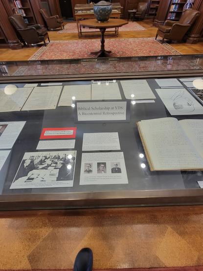 A glass vitrine in a library with dark wood and leather wing-backed chairs. A paper sign in the vitrine reads "Biblical Scholarship at YDS: A Bicentennial Retrospective. Numerous papers and photos are visible.