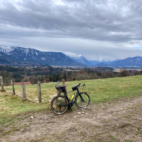 ein Fahrrad lehnt an einem Weidezaun, davor ist ein Pfad durch die Wiese erkennbar. Im Hintergrund geht der Blick über eine Ebene in Richtun