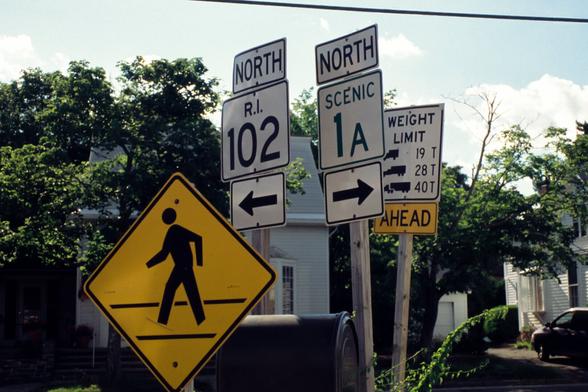 A color photograph of a number of road signs at an intersection. One reads “North - R.I. 102” and points left.  Beside it, a second sign reads “North - Scenic 1a” and points to the right.