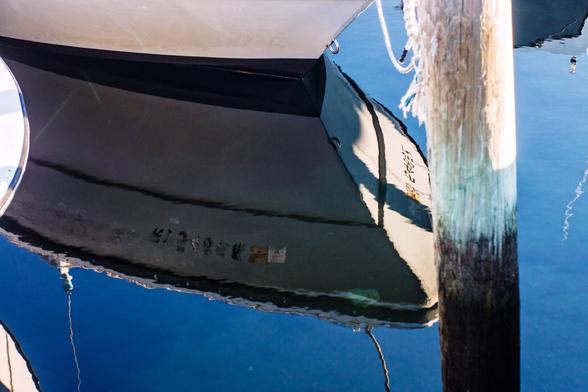 A color photograph of the bow of a small boat moored to an adjacent wooden piling, focusing on the waterline. Only the first foot or so are visible above, but the entire bow, the piling, and the sky above are clearly reflected in the blue water.