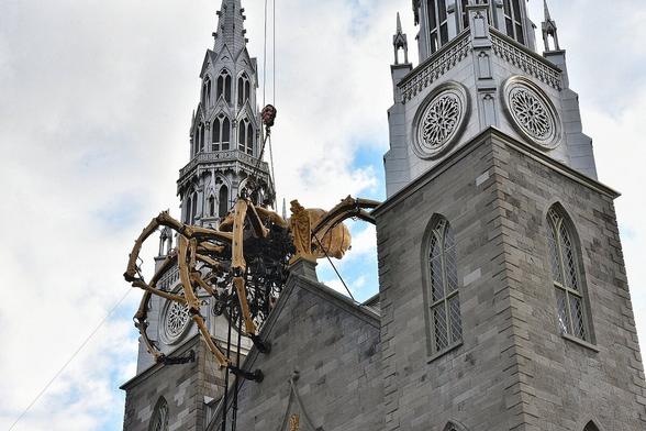 Kumo, a giant mechanical spider, is positioned between the spires of Ottawa's Notre Dame Cathedral. Kumo is part of the French production company, La Machine.

Cables that support the apparatus can be seen stretching upward and diagonally, toward the bottom left. Clouds fill the sky but some blue is visible, too.