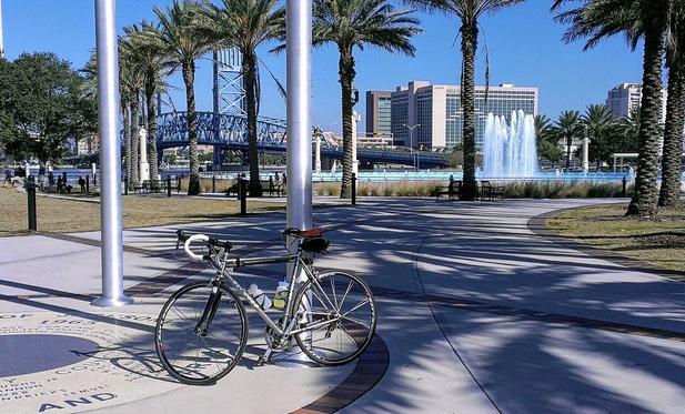 A road bike resting against a pole in a community park with fountains and a steel bridge in the distance.