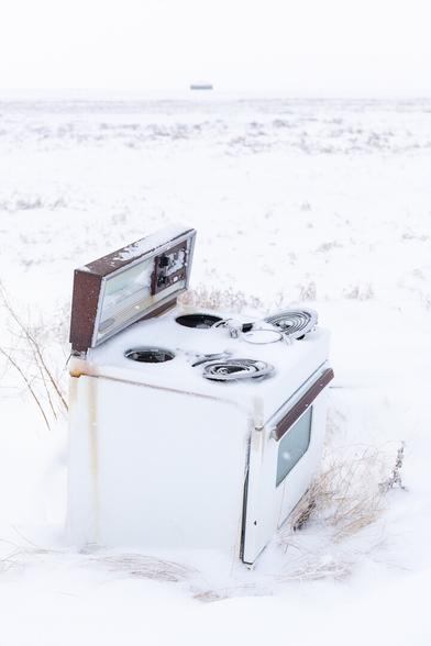 An old 1960s electric enamelled cooker (oven with hob) stands in deep snow, miles from anywhere else, near an abandoned farm. Its hob rings are bent and its starting to rust