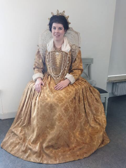 A woman sitting on a chair dresses in a golden dress, 14th century style. In her hair she wears a golden headband. Around the neck necklaces in gold and with pearls.