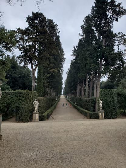 The view is up a a gently rising cypress lane. The trees are mature, with finely clipped hedges on the side, obscuring their trunks. There are statues on either side of the entrance, and one cam see statues in the classical style at regular intervals up the lane. A group of 3 people are eambolg up the lane in the distance. The lane itself is made of grit.