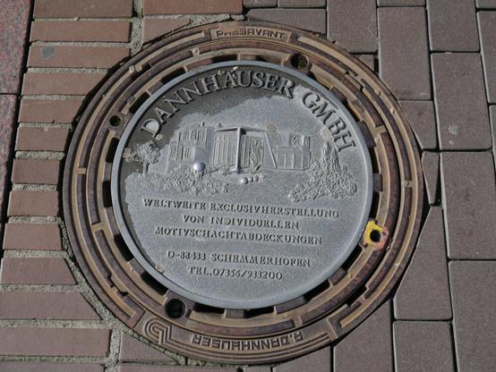 Ein Kanaldeckel mit der eingegossenen Werbung "Dannhäuser GMBH - Weltweite Exclusiverstellung von individuellen Motivschachtabdeckungen, D-33433 Schemmerhofen, Tel. 07356/93320"