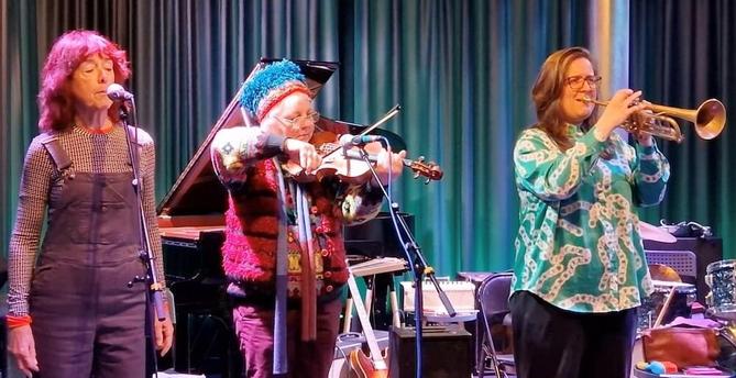 Viv Corringham and fellow artists improvising music with voice, Violin and trumpet on stage, in the background is a grand piano and an electric guitar and amplifiers. Announcement for mopomoso tv, all info in posting.