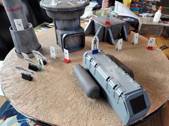 Desert terrain with paper miniatures and futuristic looking buildings and vehicles.
