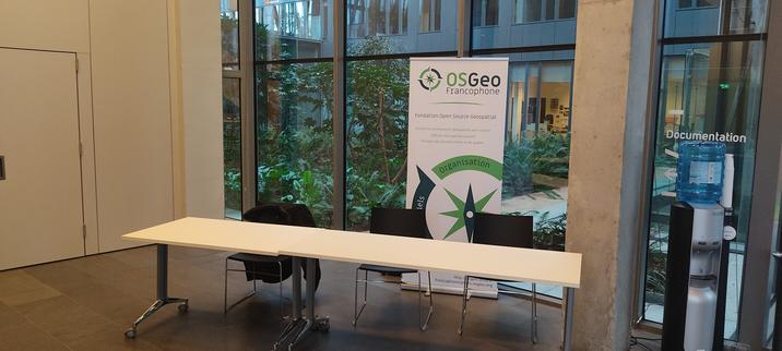 Une table et trois chaises placées dans un hall d'accueil. Derrière est placé un kakémono de l'association OSGeo-FR. L'arrière plan est un jardin arboré