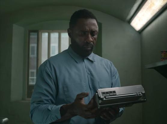 A charismatic, tough, but dejected looking early middle-aged black man stands in a prison cell holding a portable radio. He wears a blue longsleeve shirt buttoned only at the collar. He has short black hair and a goatee. A dirty fluorescent light shines above him to his left, and a small window with bars on it is behind him.