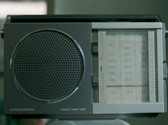 Closeup of a Grundig Party Boy 100 portable radio. It is primarily shades of grey and has a single large speaker on the left, a carrying handle on top, and a large analog readout for tuning stations that stretches from top to bottom and takes up the remaining side of the device to the right of the speaker. An orange physical tuning needle is tuned to FM frequency 65.8.