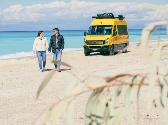 Steffi und Lui spazieren an einem weißen Sandstrand. Im Hintergrund steht ihr knallgelber Camper.