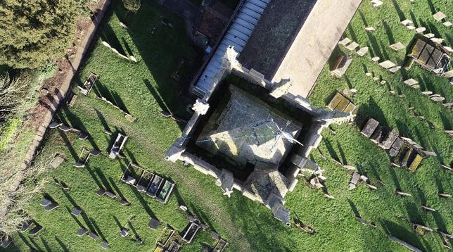 Vertical view down on to a square church tower. Eight pinnacles on crenellations. Central pinnacle. Below the graveyard with rows of headstones, all casting shadows in bright sunlight. The nave roof runs out of view top centre right.