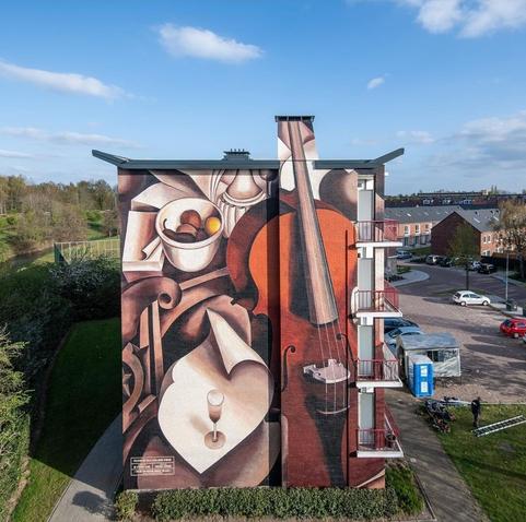 Streetartwall. On the outer wall of a modern, four-storey and detached building, an impressive mural of a still life with string instrument is sprayed / painted. The image is kept in brown tones , beige and white. Some sheets of paper, a glass of wine, a small bowl of fruit, and on the right a large cello (or is it a violin, sorry) whose neck protrudes and was painted on the chimney.