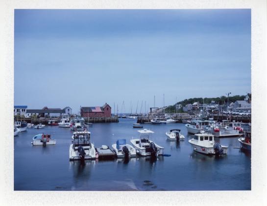 A white bordered, rectangular color Polaroid photograph of a red shack on a pier.  A harbor surrounded by blue water and skies, and moored boats. There’s an American flag on part  of the roof.

The shack is the replica of the original 1840 fisherman’s shack built in 1840. Rockport, Massachusetts felt it was so iconic that they rebuilt it after it was destroyed in the Blizzard of 1978.