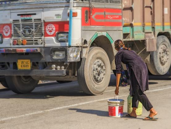 A photo of a girl carrying two buckets across a highway, waiting for a big truck to pass