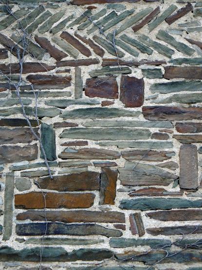 Color photo of a portion of a house wall featuring irregular courses of brown and green stones of various thicknesses and lengths set into white mortar
