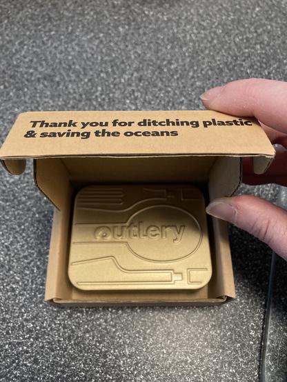 A hand opening a box with a small metal tin inside that says “Outlery” on it