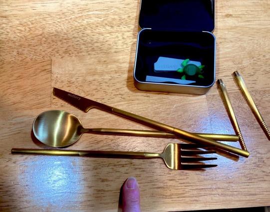 A set of gold toned silverware and a small tin the silverware came out of, on a wooden table