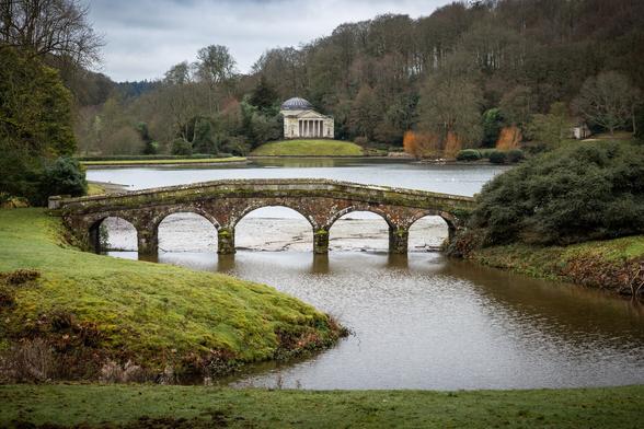 Stourhead National Trust in Wiltshire, England.  Old brick bridge over a lake surrounded by woodland and old building in the distance beyond the bridge and lake