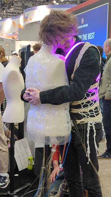 A person hugging a mannequin. The person is wearing a harness of wires around their body, to simulate the feeling of being hugged.