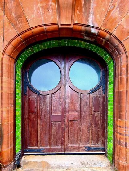 A pair of wooden doors double in a red sandstone doorway with a smoothly curved top outlined in green tiles.