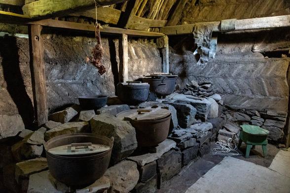 Inside a room in an old wooden building. It's dark, with dark wooden beams on show and the hint of some form of dirt or peat brick panelling in the walls. Some stone blocks form a trough of sorts and some large metal pots are resting in sectioned off areas of the trough. It looks like some antiquated form of multiple hobs in a fireplace. It feels enclosed and quiet here, but protected from the elements.