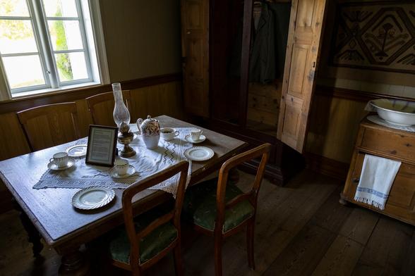 Inside a room in an old wooden building. The walls are panelled in wood and there is furniture in keeping with a dining room - cupboards and chests of drawers, plus a table with four chairs set at it, two against each of the slightly longer lengths. Behind the table is a window letting in light to this place. In contrast to the last photo, this feels more modern, but still lacking much in the way of modernity. The table is set with some plates, cups and saucers, and an oil lamp.