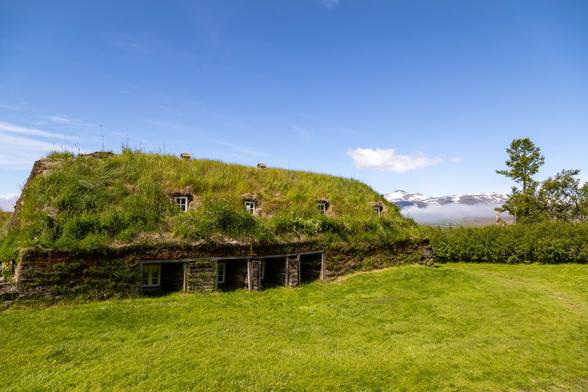 Outdoors, on a bright, sunny day with mostly blue sky. We're standing on lush, green grass at the rear of a building built from thick stone and roofed with grass. There are four recesses into the wall at the rear in which some small windows are set almost at ground level, and in the roof space four more windows can be seen amongst the overgrown grass, although not in line with the windows below. There's a feeling that the house has evolved and had changes applied over time, and with the roll and rise and fall of the grass we're standing on we can tell that the house is partially submerged into the landscape for protection from the elements. Just beyond the building in the distance to the right we can see a low bank on cloud or mist on the horizon obscuring the base of a snow-peaked mountain range. It feels very peaceful here.