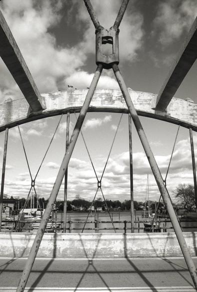 A black and white photograph of a view though a concrete rainbow arch bridge, showing the cove behind it. The picture was taken mid span, perpendicular to the roadway, for the opposite side fro. The cove. The far side arch, the overhead concrete members linking the arches, and the metal supports on the near and far side are all visible, as is some of the roadway.