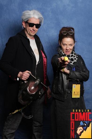 Peter Nolan in cosplay as the Twelfth Doctor (Magician's Apprentice outfit) next to Michelle Gomez as she points a banana at the camera like a gun.