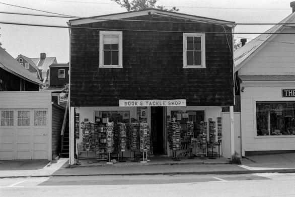 A black and white photograph of a somewhat ramshackle commercial building in a seaside community. The building is on an incline and appears slightly crooked. The  shingles on the overhanging top story are stained a dark color and two windows look out over the street. The ground floor is painted white. Under the overhang, eight or nine spinning  display racks of postcards are  clumped together. Over the door, a hand painted sign reads “BOOK & TACKLE SHOP”.