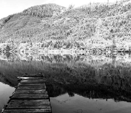 A wooden dock is seen on lower left of image.  Lake with mountains in background. Mountains are brightly lit by sunlight. Black and white photo.