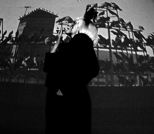 Lily Fawn, with a crow on her head & a SFX bloody bird pecking wound on her face, standing in front of film projection of a bunch crows from Alfred Hitchcock's - The Birds - movie & holding a cigarette near her mouth.