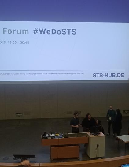 An auditorium hall. On the wall there is a slide that shows the hashtag #WeDoSTS