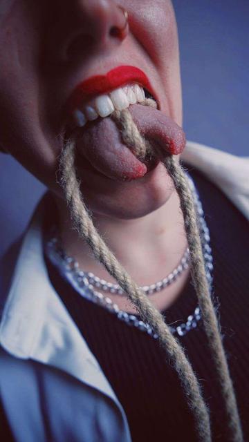A person wearing red lipstick, a black undershirt, white shirt, silver chains, and a 1/4 inch hemp rope tied tightly around their split tongue.