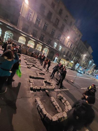 Scène de nuit, sur une place, des gens disposent des pavés de manière à composer FUCK 49.3 !