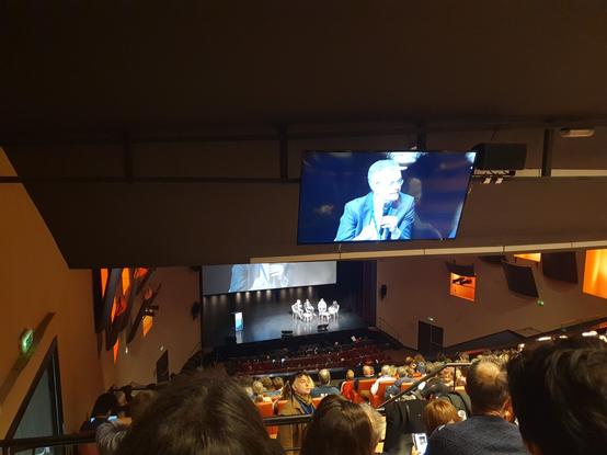 Quatre personnes assises sur scène en arrière-plan et reprise vidéo sur Thomas Fatome